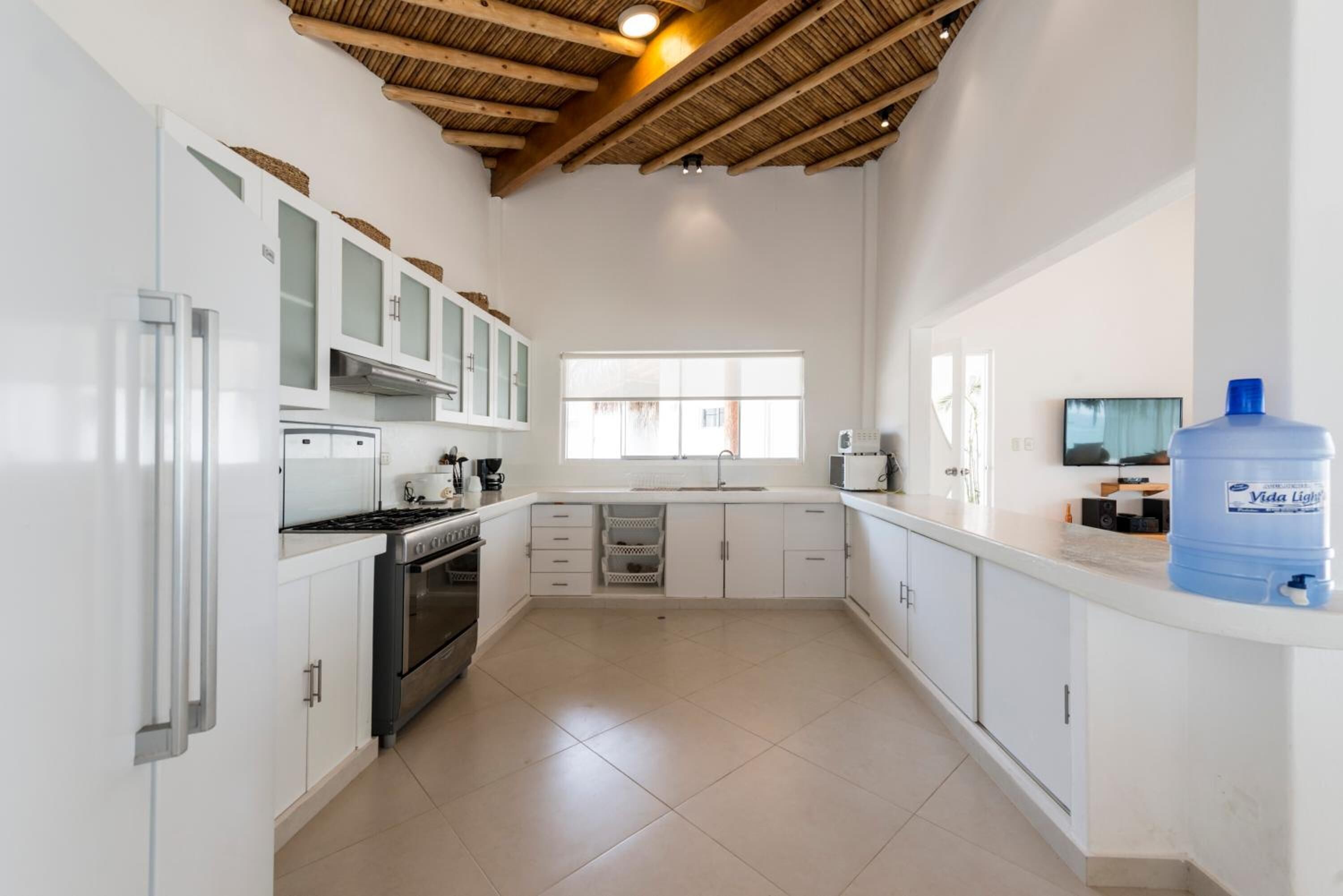 Una cocina moderna con armarios blancos, electrodomésticos de acero inoxidable, un dispensador de agua grande y un ventanal que proporciona luz natural.