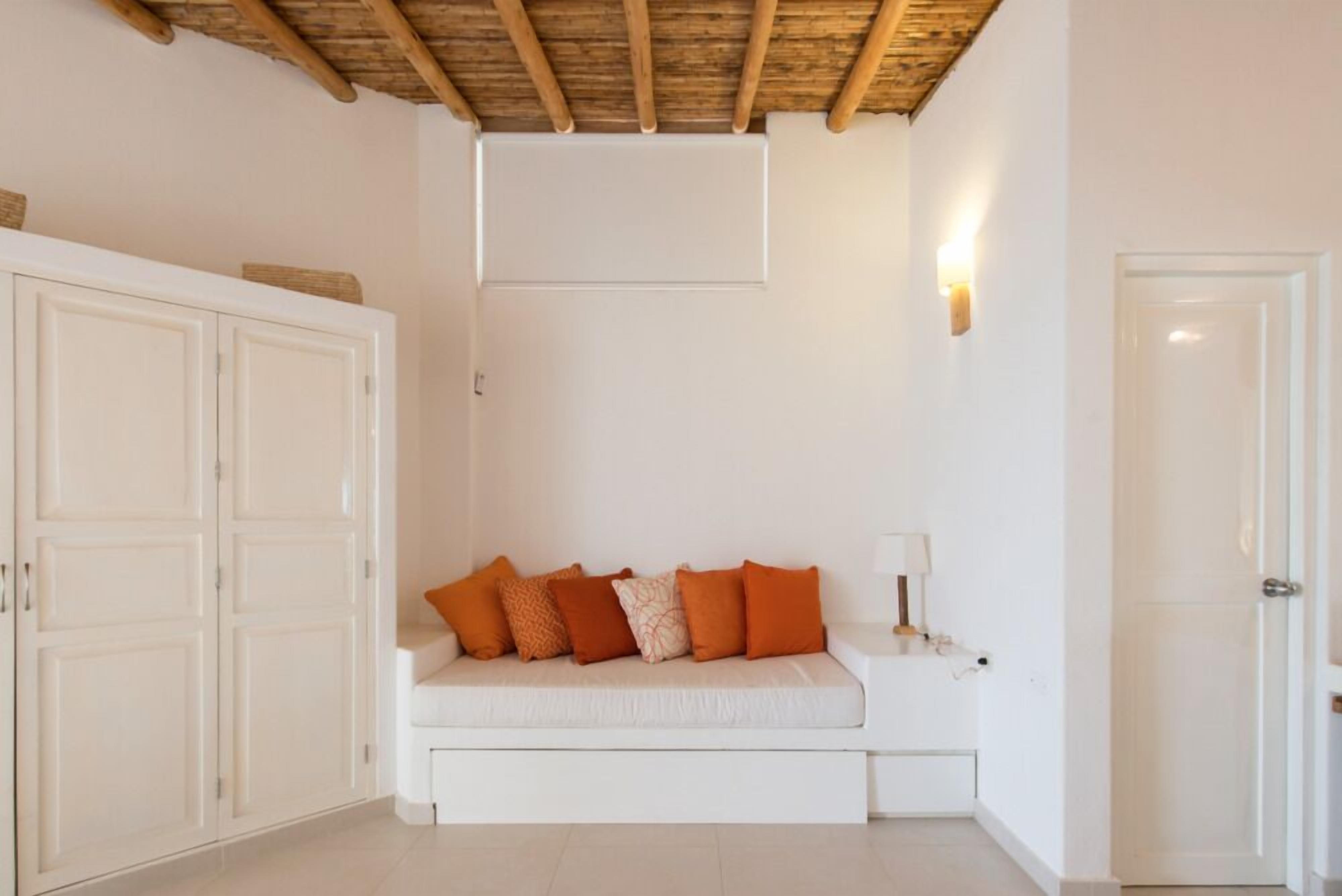 A room with a white sofa, orange cushions, a wooden ceiling, a white wardrobe, and a door.