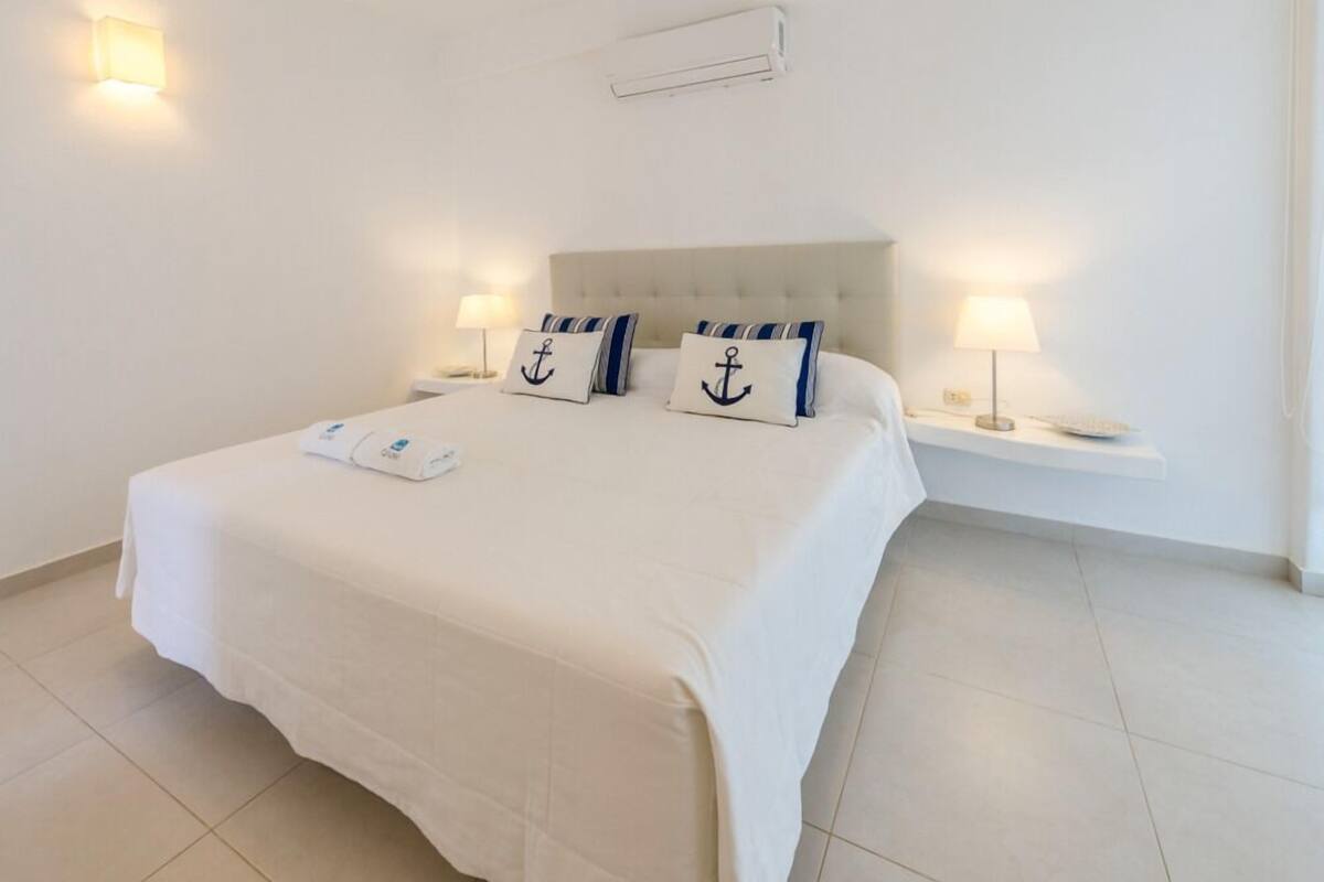 A neatly made bed with a white bedspread and two pillows with anchor designs, flanked by two bedside lamps on white nightstands.