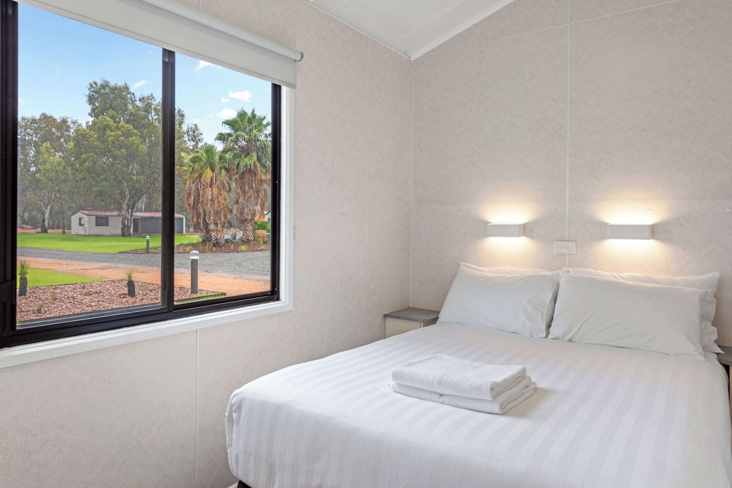 A hotel room with a large window, a neatly made bed, and two wall-mounted lights.