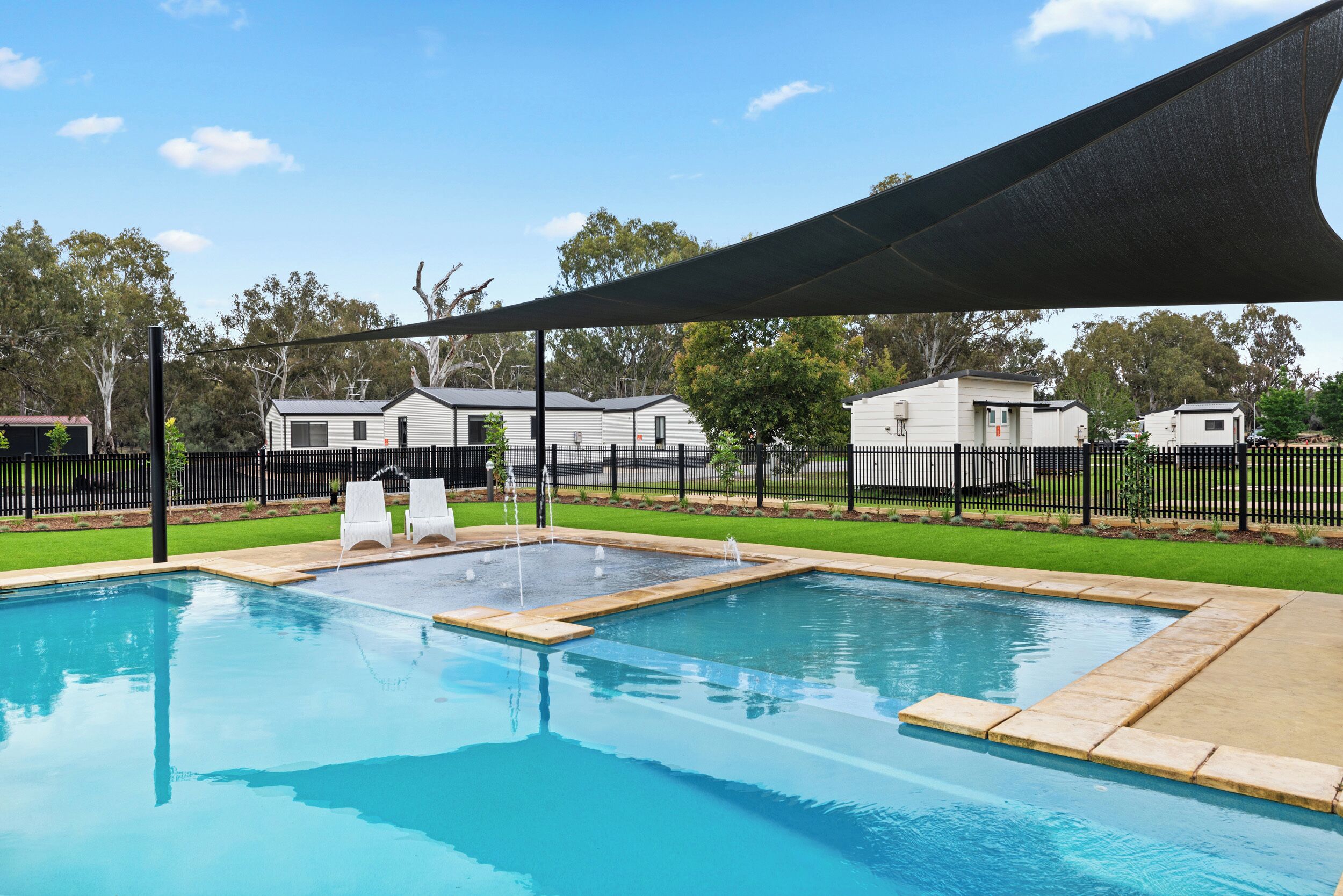 Une piscine avec deux chaises longues, un espace ombragé et une clôture entourant un quartier résidentiel avec des maisons.