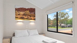 A hotel room with a large bed, a nightstand, a wall-mounted light, and a window with a view of a park and buildings. - Discovery Parks - Mildura (Buronga)