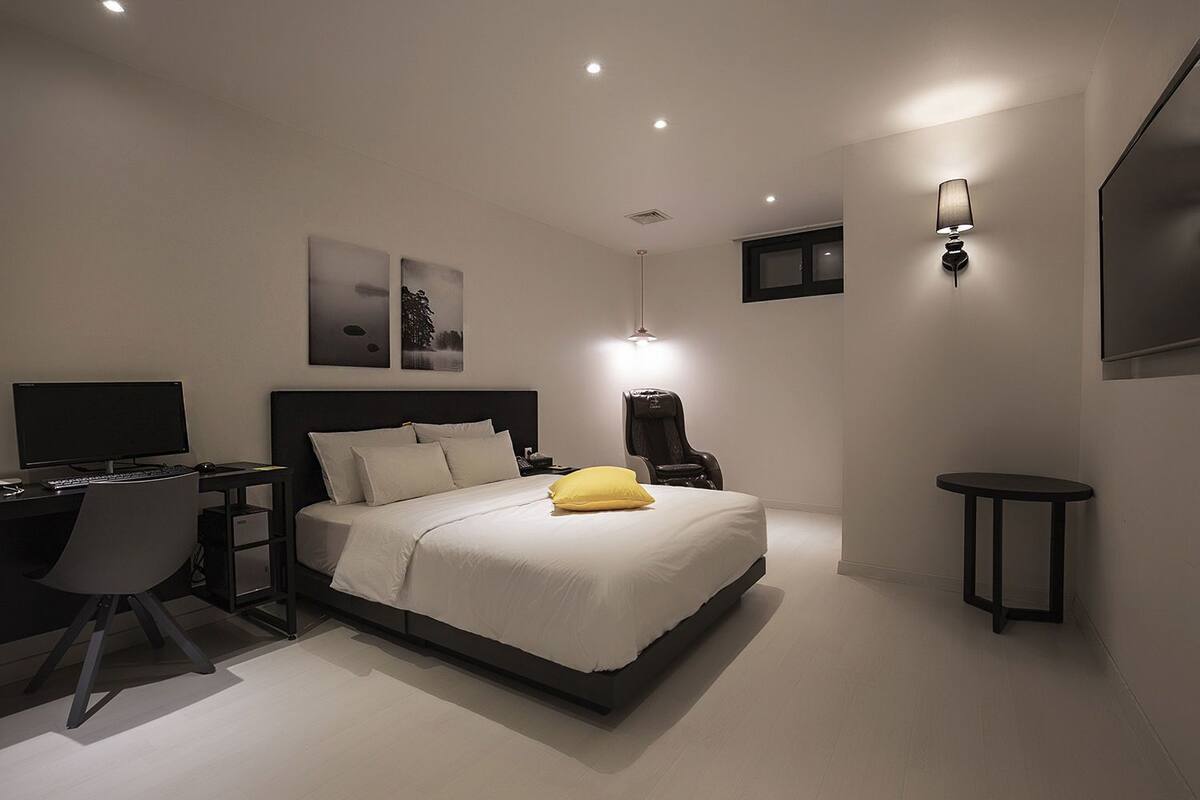 A modern bedroom with a bed, a desk, a chair, a lamp, and artwork on the wall.