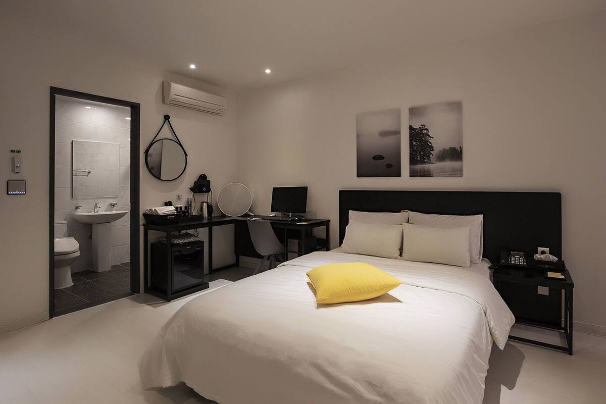 A hotel room with a bed, a desk, a chair, a computer, a phone, and a nightstand.