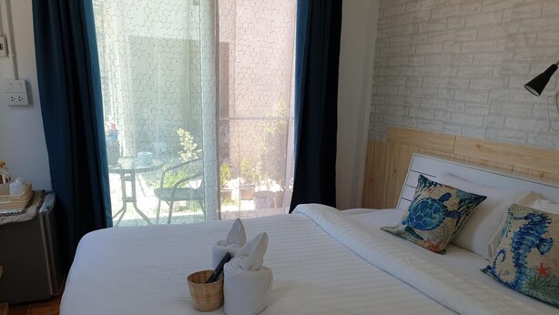 Una cama bien hecha con ropa blanca, cabecera y almohadones decorativos. Hay una cesta de mimbre con una toalla y una botella al lado.