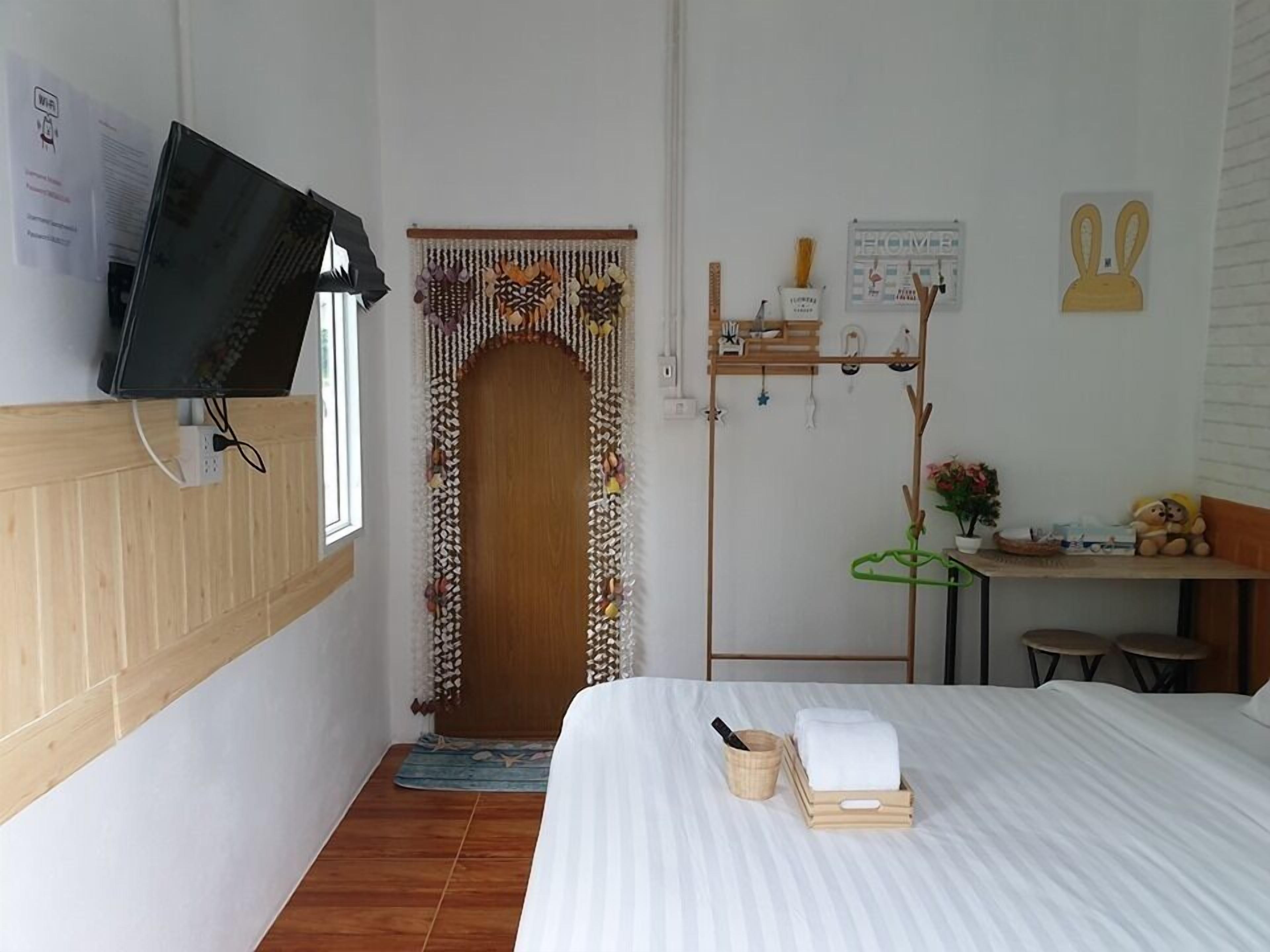 Une chambre d’hôtel avec un lit, un téléviseur fixé au mur, une porte en bois agrémentée d’un rideau décoratif, une petite table avec une plante et un porte-serviettes mural avec des serviettes et des articles de toilette.