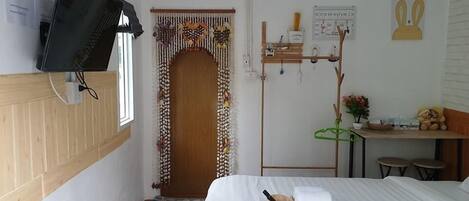 Une chambre d’hôtel avec un lit, un téléviseur fixé au mur, une porte en bois agrémentée d’un rideau décoratif, une petite table avec une plante et un porte-serviettes mural avec des serviettes et des articles de toilette.