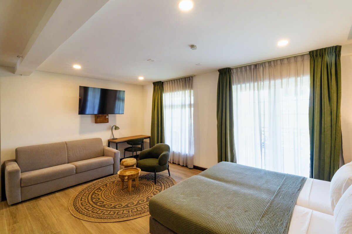 Una habitación de hotel moderna con sofá, sillón, cama, televisor y escritorio.