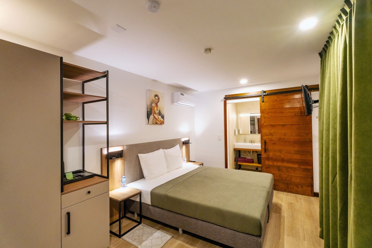 Un dormitorio con una cama, un escritorio, una estantería, un cuadro y un baño visible a través de una puerta abierta.