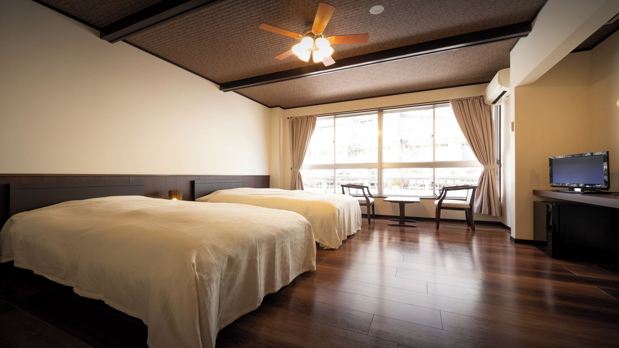 A hotel room with two beds, a ceiling fan, a TV, a table, and a window with curtains.