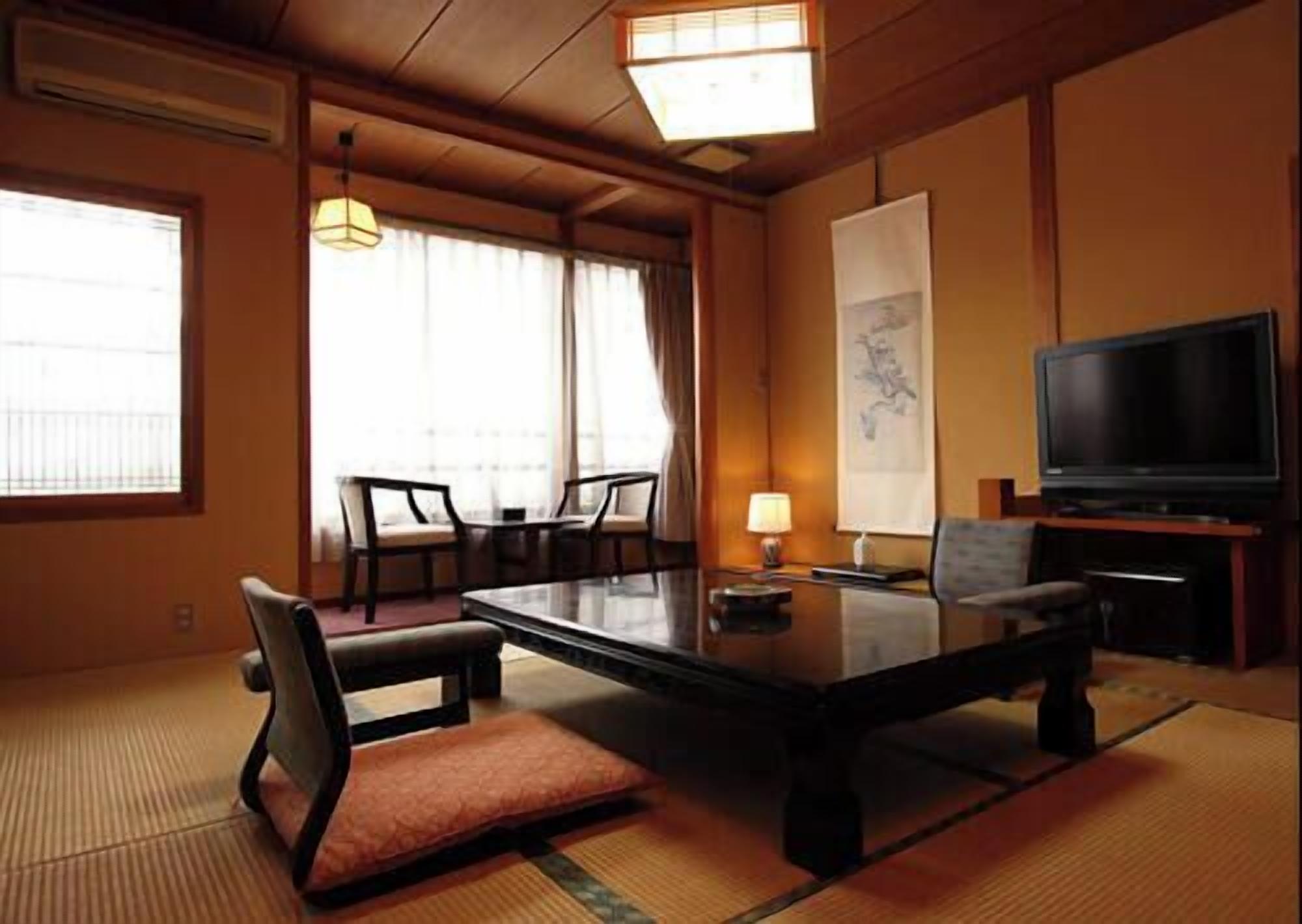 A hotel room with tatami flooring, a low table, chairs, a television, and a large window with curtains.
