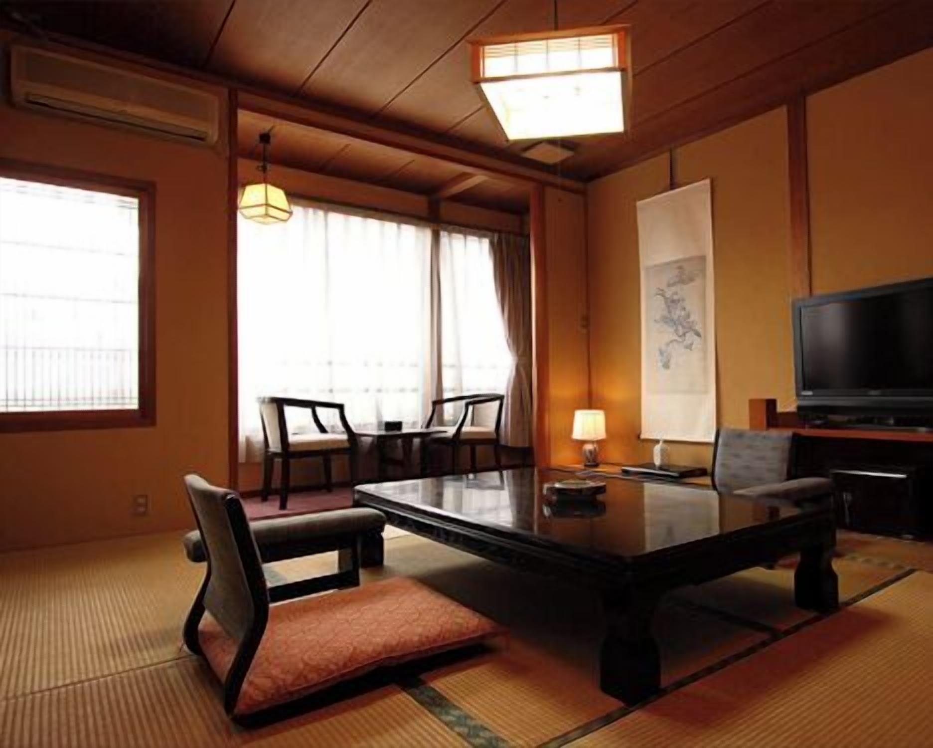 A traditional Japanese-style room with tatami flooring, a low table, chairs, a television, and a large window with curtains.