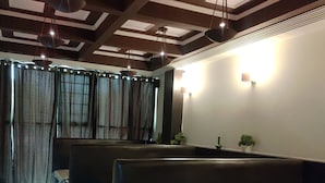 Ein Zimmer mit niedriger Decke, dunklen Holzbalken und einem großen Fenster mit gestreiften Vorhängen. Zwei schwarze, gepolsterte Sitzbereiche und ein kleiner Tisch mit einer Plastikdecke sind vorhanden.