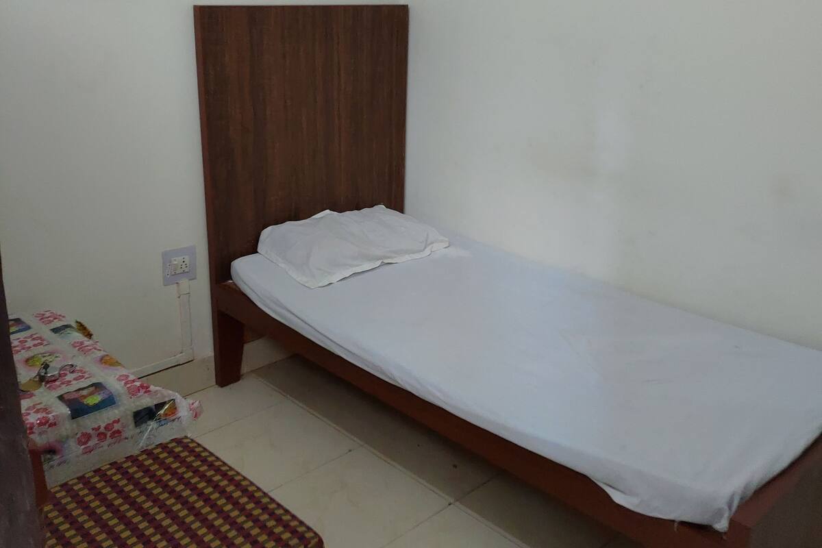 A small, single-bed room with a wooden headboard, a bedside table, and a bench with a patterned cushion.