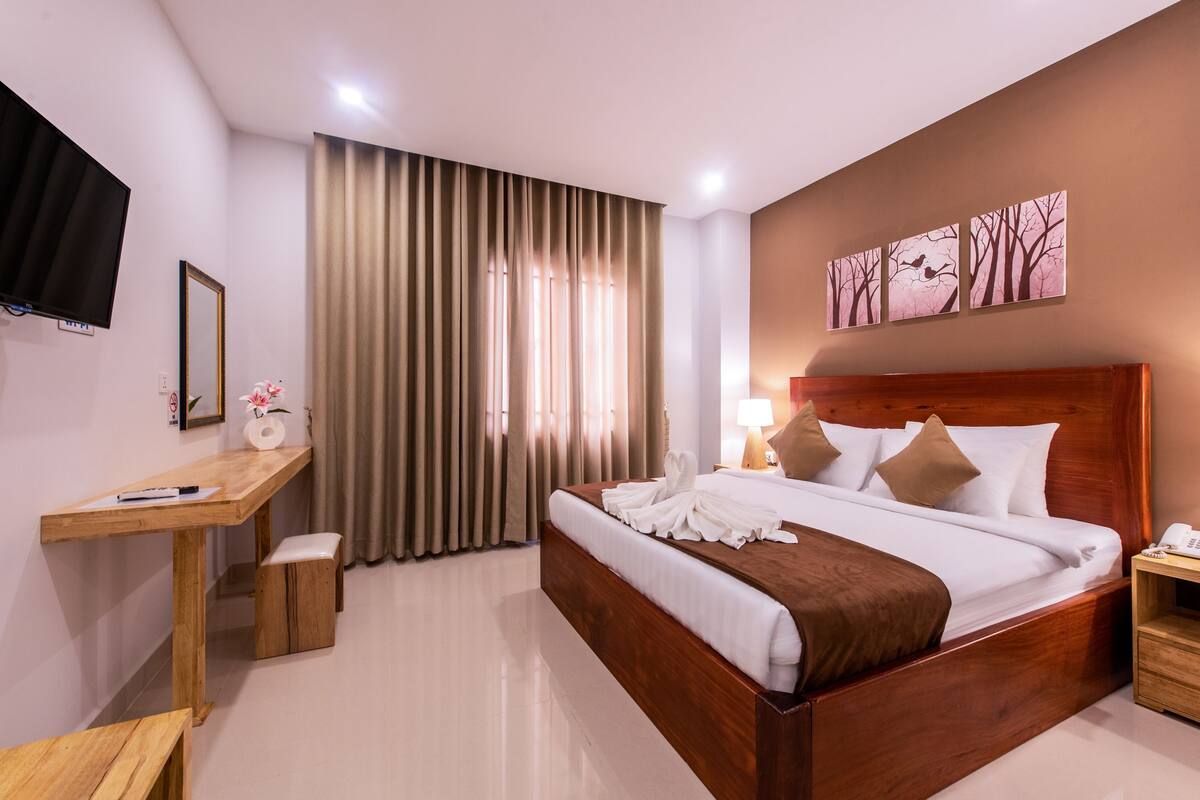 Une chambre d’hôtel avec un lit en bois, un bureau, une chaise, une télévision et une fenêtre avec des rideaux.