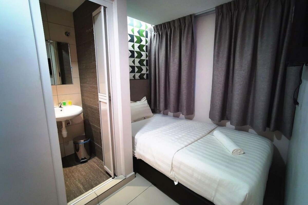 Une chambre à coucher avec un lit, des rideaux, une fenêtre ornée d’un motif géométrique, et une salle de bain avec un lavabo et un miroir.