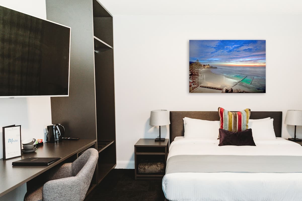 Ein Hotelzimmer mit einem Bett, zwei Nachttischen, einem Schreibtisch mit Stuhl, einem Fernseher und einem Bild mit Strandmotiv.