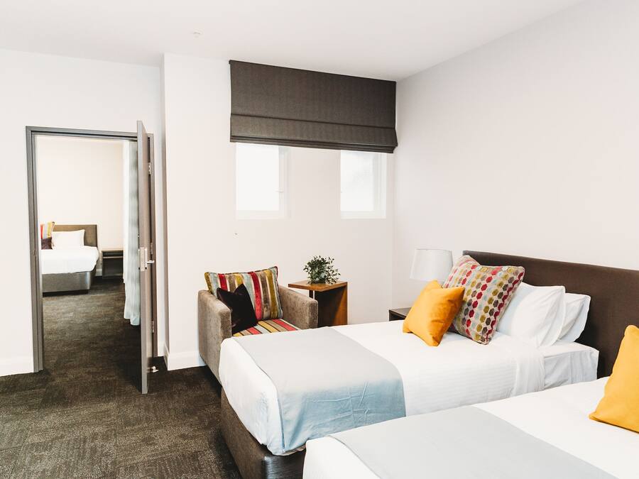 A hotel room with two beds, a sofa, a small table with a lamp, and a window with a blind.