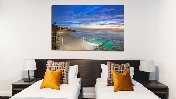 A hotel room with two beds, a large beach view painting, and a nightstand with a lamp. - Coniston Hotel Wollongong (Coniston)