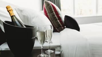 A bottle of champagne in a black bucket, two glasses of champagne, and a bed with white bedding and a striped pillow in a hotel room.