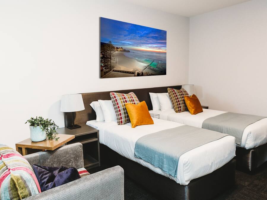 Ein Hotelzimmer mit zwei Betten, einem Nachttisch, einer Lampe, einem Sessel und einem Bild mit Strandmotiv.