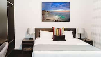 A hotel room with a large bed, two bedside tables with lamps, a nightstand, a chair, and a wall-mounted painting of a beach scene.