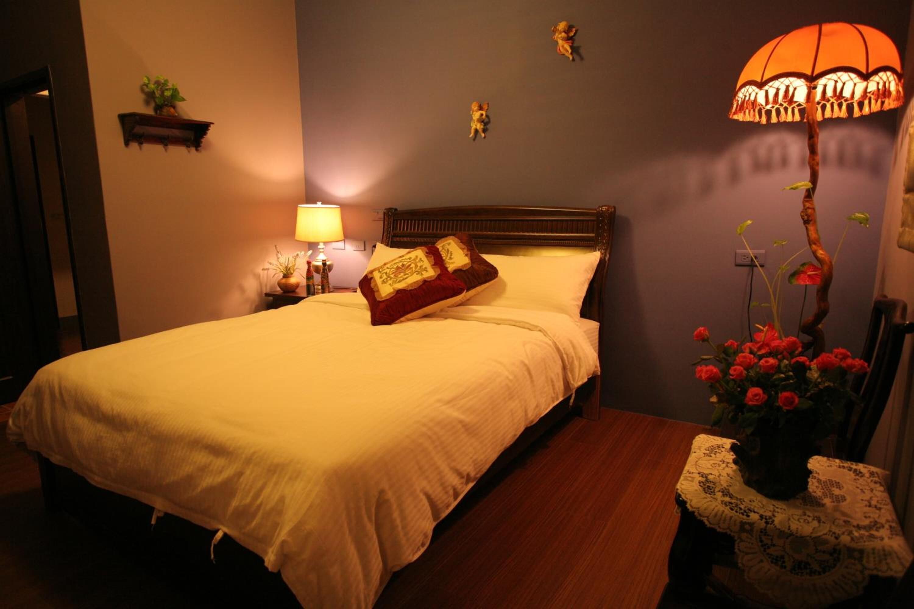 A neatly made bed with a white comforter and a red pillow, a bedside lamp, a small plant on the nightstand, and a vase with red flowers on a table beside the bed.