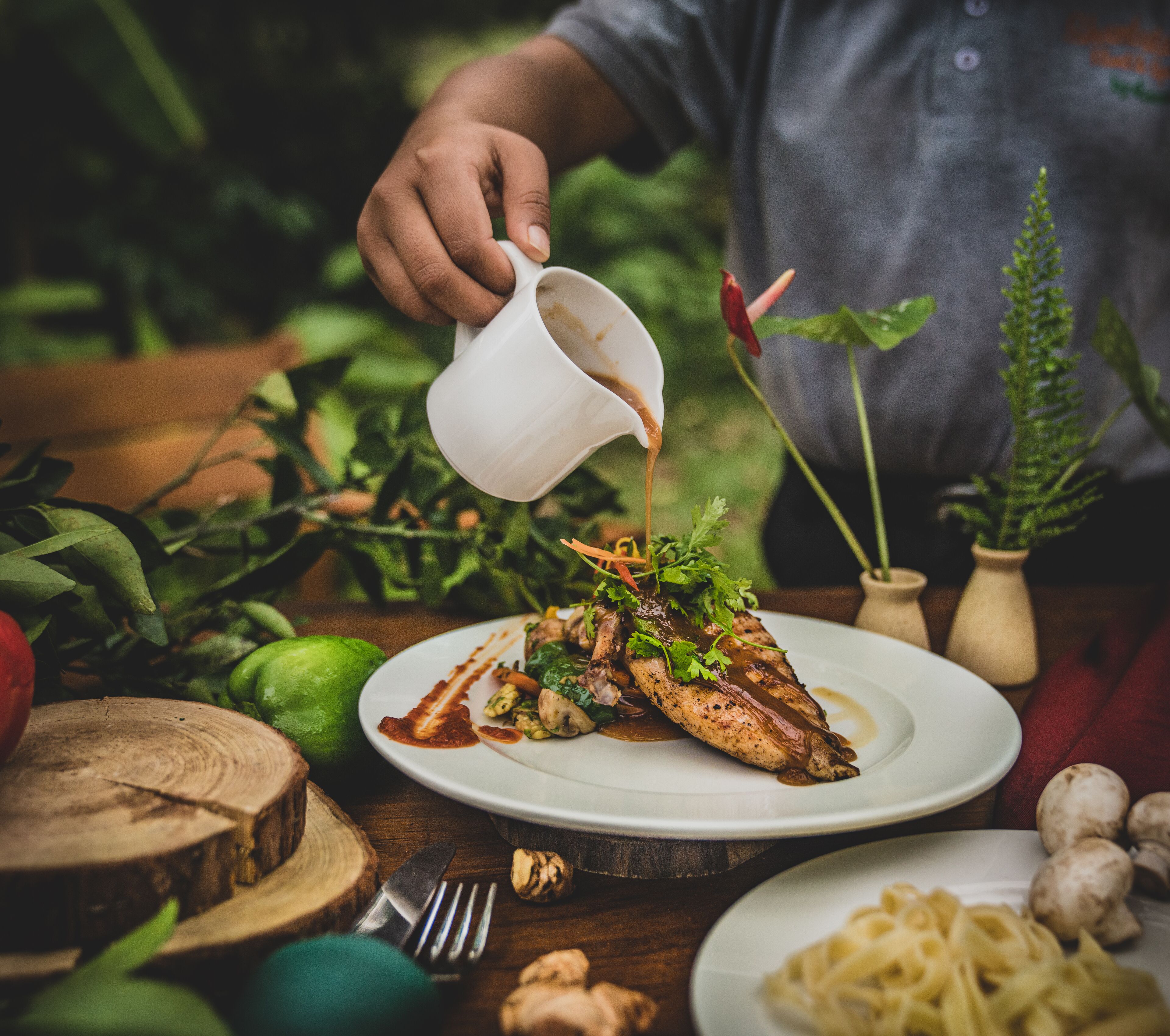 A person pouring sauce over a dish of grilled chicken and vegetables on a white plate.