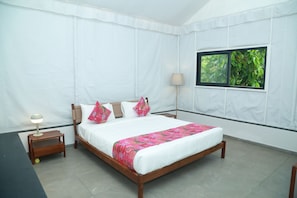A bedroom with a wooden bed, white curtains, a window with a view of greenery, a bedside table with a lamp, and a small shelf. - Shambala Yercaud (Yercaud)