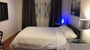 Een netjes opgemaakt bed met een wit-grijs gebeddegoed, een nachtkastje met een lamp en een afstandsbediening erop.