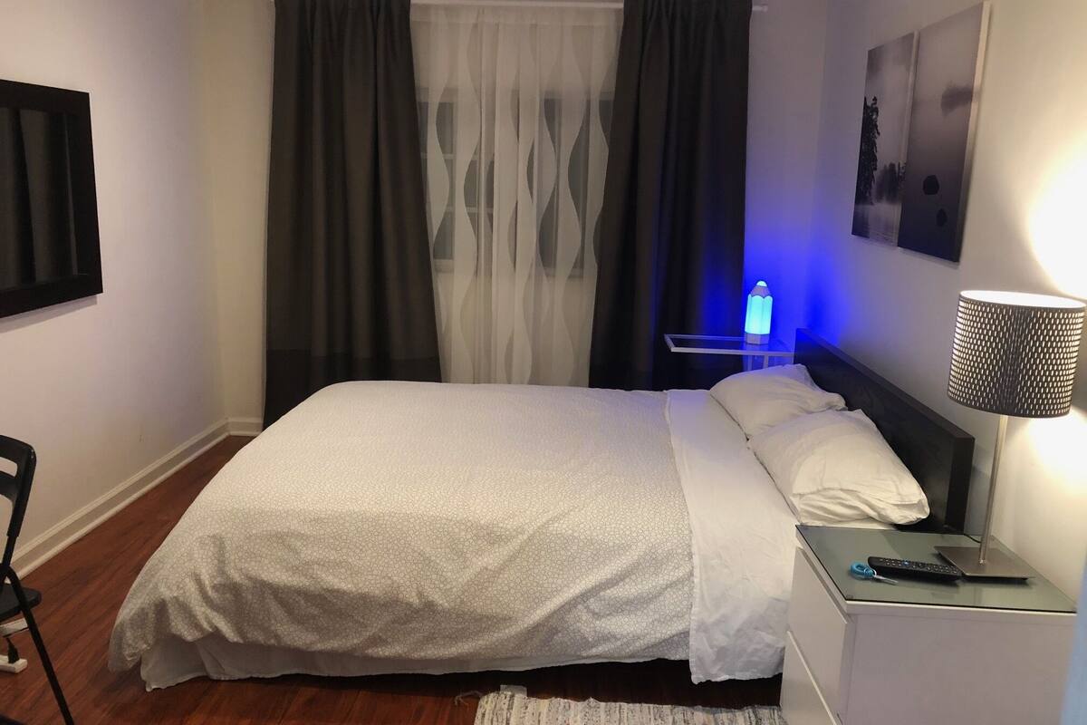 A neatly made bed with a white and grey patterned bedspread, a bedside table with a lamp, and a remote control on it.