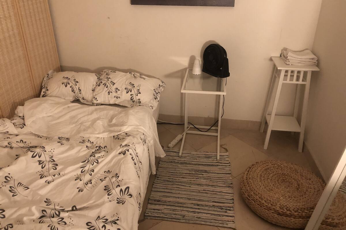 A bedroom with a bed, a small table with a hat, a woven pouf, and a painting on the wall.