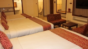 A hotel room with two large beds, a sitting area with a chair and a small table, a television mounted on the wall, and a bathroom visible through an open door. - Kaveri Hotel (Mysore)