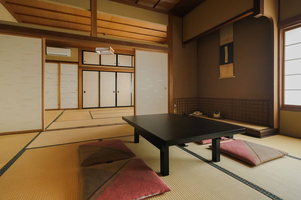Ein traditioneller japanischer Raum mit Tatami-Böden, einem niedrigen Tisch, Kissen, Schiebetüren und einem Fenster mit Ausblick.