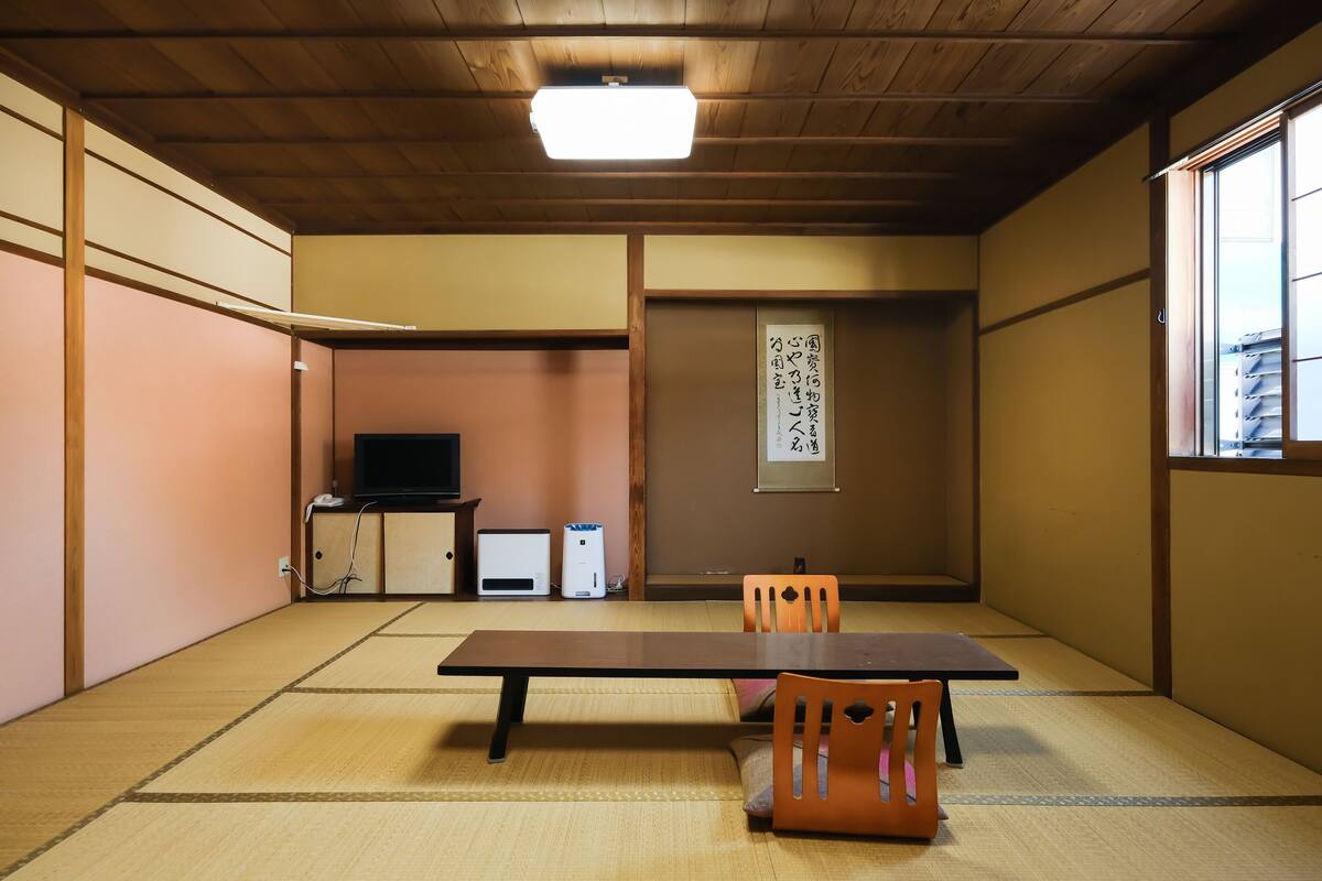 Ein traditioneller japanischer Raum mit Tatami-Böden, einem niedrigen Tisch, zwei Stühlen, einem Fernseher und einer Schiebetür.