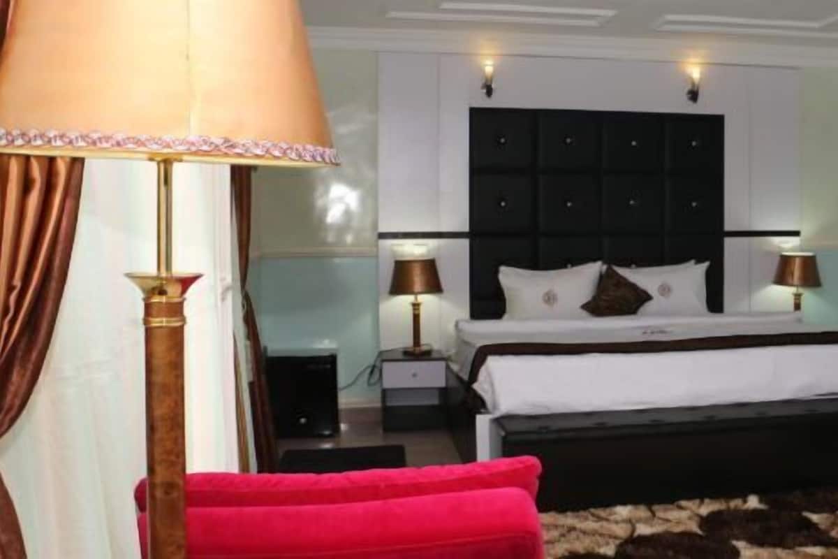Um quarto com cama, duas mesas de cabeceira, uma cadeira vermelha, luminária e um quadro na parede.