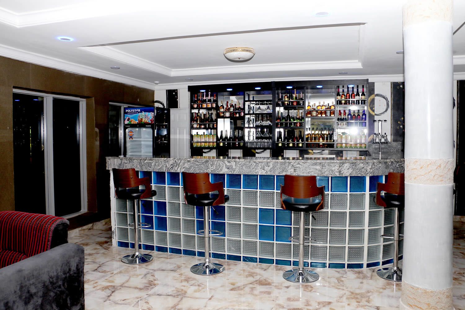 A bar area with a marble countertop, multiple stools, and a variety of bottles and glasses on shelves.