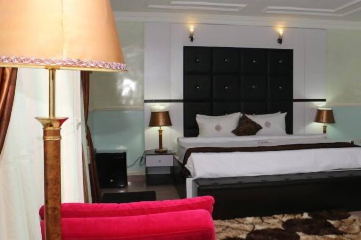 Quarto com cama, mesas de cabeceira, abajur, cadeira vermelha e tapete estampado.
