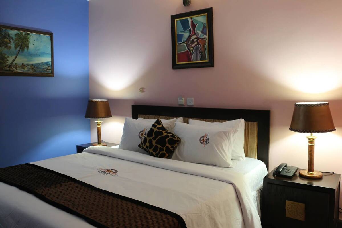 Quarto de hotel com uma cama, duas luminárias de cabeceira, mesa de cabeceira com telefone e duas pinturas penduradas na parede.
