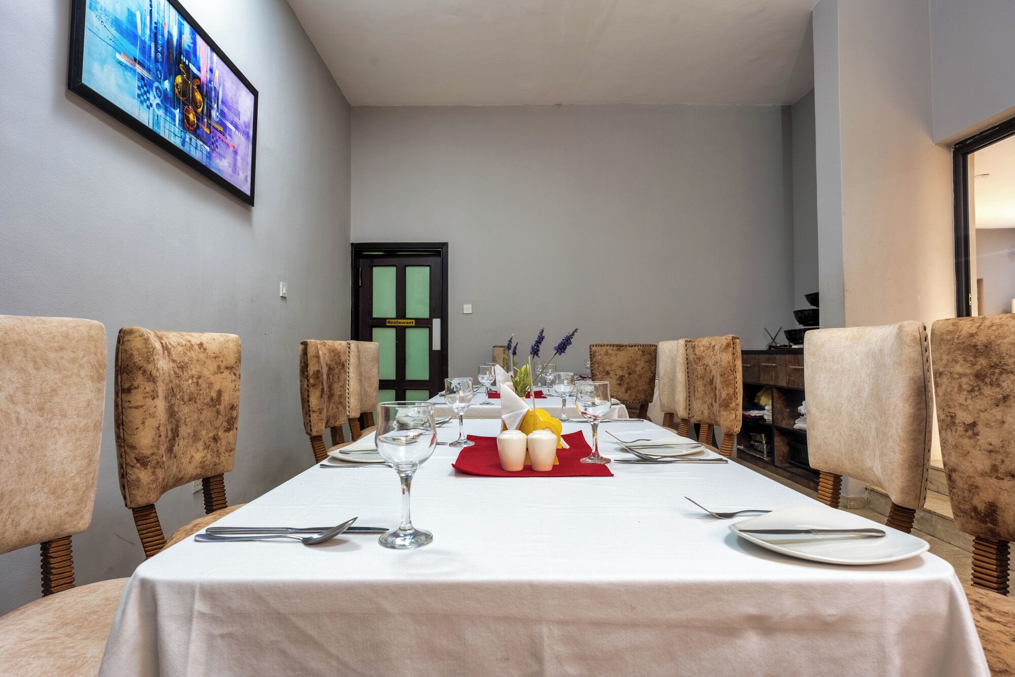 A dining room with a long table set for a meal, flanked by upholstered chairs, a framed artwork on the wall, and a door labeled "Restaurant".