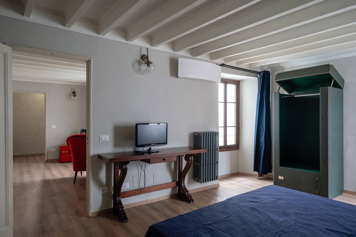 A bedroom with a bed, a desk with a computer, a radiator, and a window with curtains.