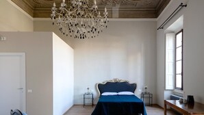 A bedroom with a large chandelier, a bed with a dark blue bedspread, two bedside tables, and a window with a view. - B&B Stupido Hotel (Florence)