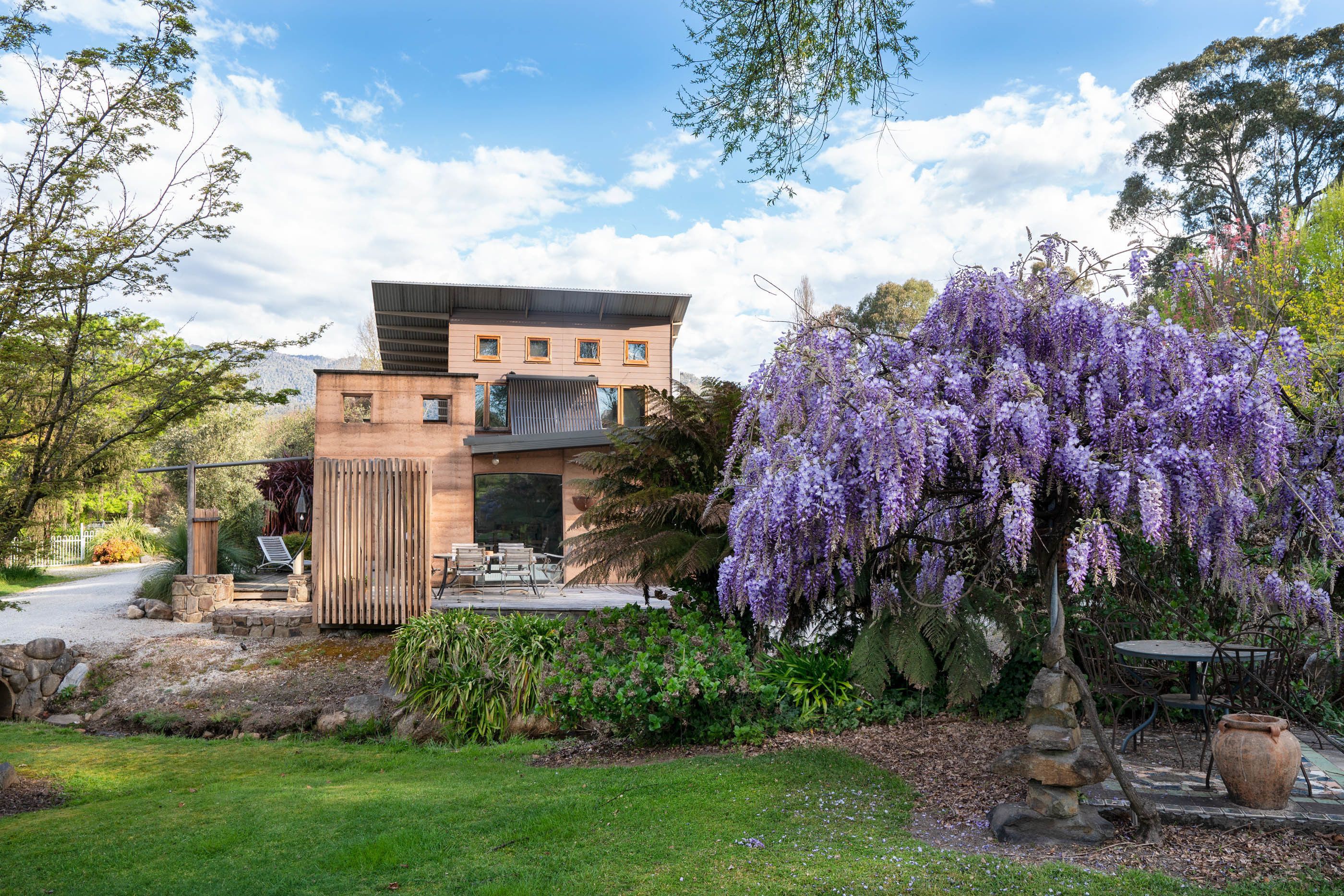 A modern house with a patio, surrounded by lush greenery and purple wisteria flowers.