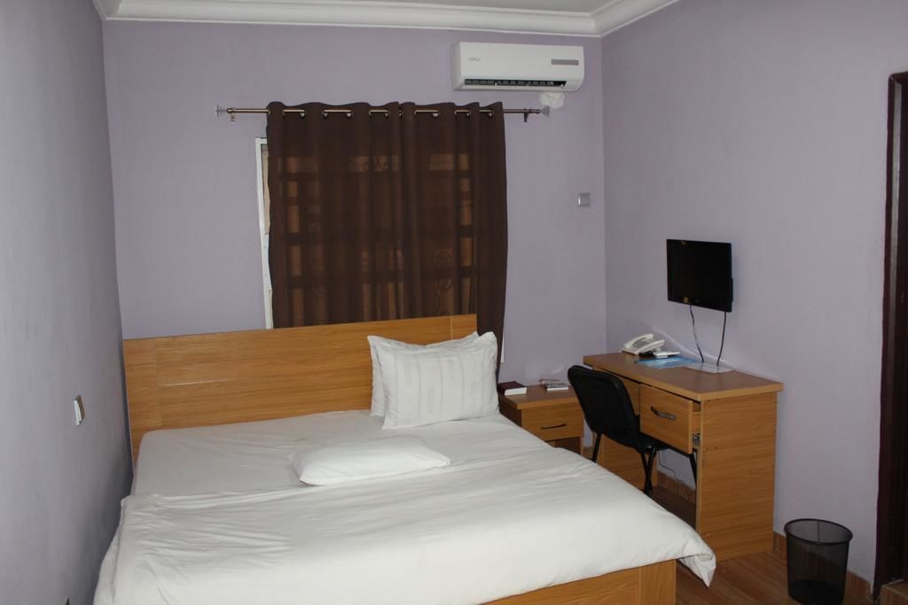 Una cama bien tendida con edredón blanco y almohadas, un escritorio de madera con una silla, un altavoz negro y un pequeño cesto de basura.