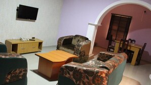 A living room with a sofa, a coffee table, a TV mounted on the wall, a wooden cabinet, and a window with blinds. - Ayoluyem Hotel (Port Harcourt)