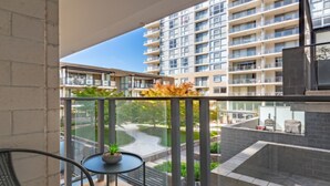 A balcony with a view of a modern apartment building, a small table with a plant, and a chair. - Spacious 1-Bedroom Apartment With Parking (Canberra)