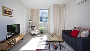 A modern living room with a flat-screen TV, a sofa, a coffee table, and a large window with curtains. - Spacious 1-Bedroom Apartment With Parking (Canberra)