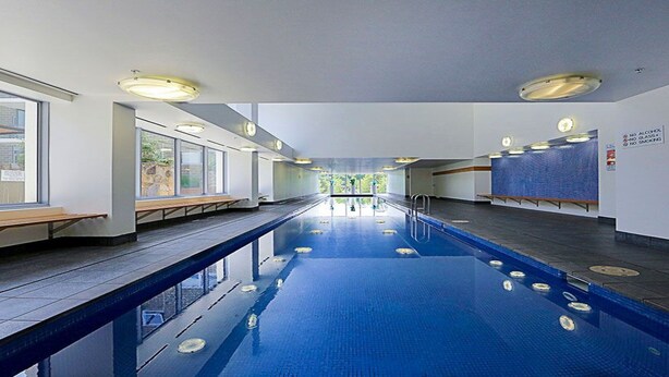 A long indoor swimming pool with clear lanes, benches along the sides, and a view of the outdoors through large windows. - Spacious 1-Bedroom Apartment With Parking (Canberra)