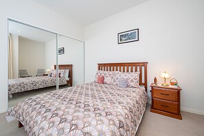 A bedroom with a large mirror, a bed with patterned bedding, a bedside table with a lamp, and a framed picture on the wall.