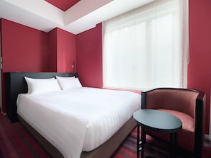 Een hotelkamer met een groot bed, een stoel, een tafeltje en een raam met dunne gordijnen.